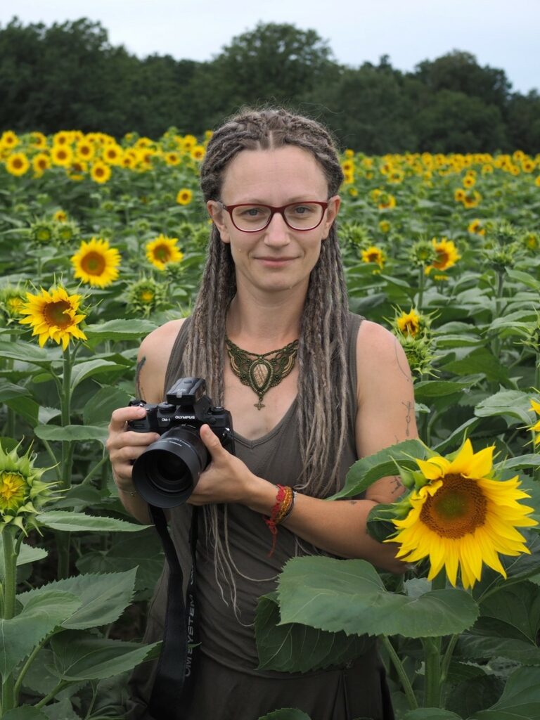 Fotografka Lenka Jamnická stojí s fotoaparátem v poli se slunečnicemi.